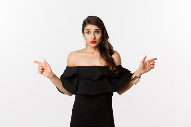 Fashion and beauty. Indecisive young woman in glamour black dress pointing fingers sideways, showing two choices and looking confused, white background.