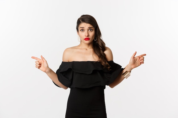 Fashion and beauty. Indecisive young woman in glamour black dress pointing fingers sideways, showing two choices and looking confused, white background