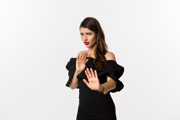 Fashion and beauty. Reluctant and worried woman asking to stay away, showing stop gesture and looking scared, standing in black dress over white background.