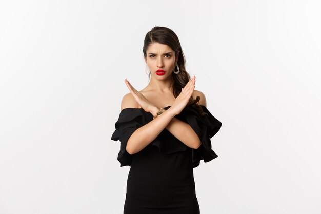 Fashion and beauty. Serious and confident female model in black dress, showing cross sign and frowning, stop gesture, telling no, standing over white background.