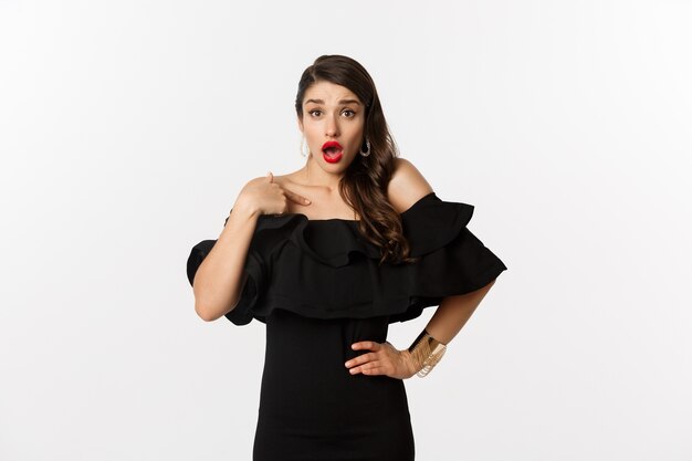 Fashion and beauty. Shocked woman pointing at herself and looking at camera, being accused, standing in black dress over white background.