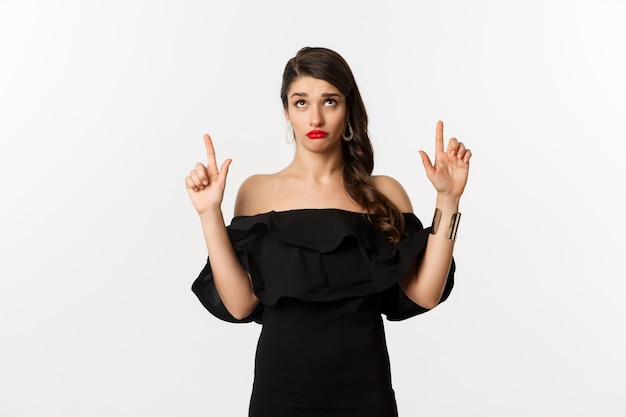 Fashion and beauty. Silly woman in black dress, red lips, looking and pointing fingers up with unamused doubtful expression, white background.