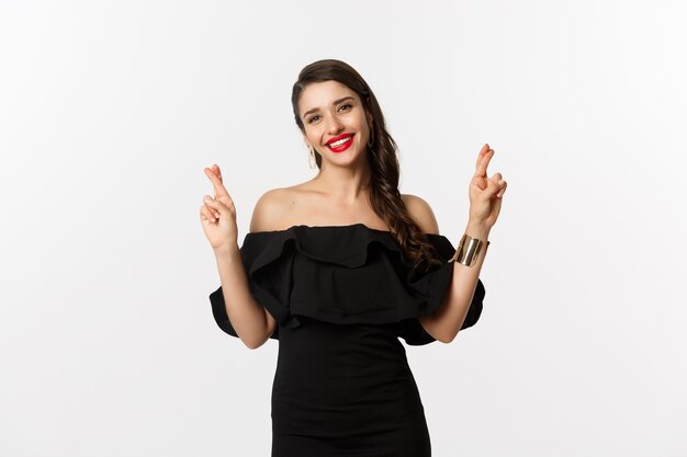 Fashion and beauty. Stylish glamour woman in black dress, red lips, looking optimistic and smiling while cross fingers, making wish, standing over white background.
