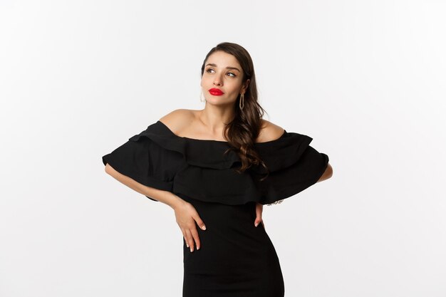 Fashion and beauty. Stylish modern woman in black dress, makeup and red lips, posing over white background self-assured, standing over white background