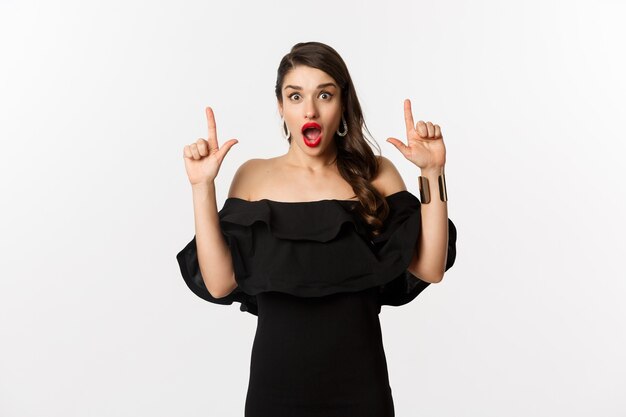 Fashion and beauty. Surprised woman in black dress pointing fingers up, showing banner, standing over white background