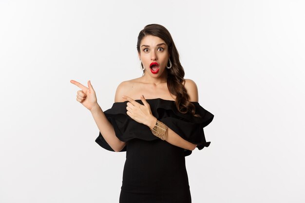 Мода и красота. Удивленная женщина в черном гламурном платье показывает пальцем влево, демонстрирует рекламу и смотрит изумленно, белый фон