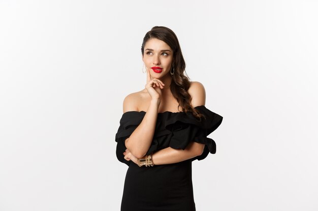 Fashion and beauty. Thoughtful young woman in black dress, smiling pleased and thinking, having an idea, white background.