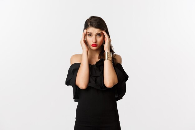 Fashion and beauty. Young modern woman in glamour dress, jewelry and makeup, touching head and looking exhausted, feeling dizzy, standing over white background