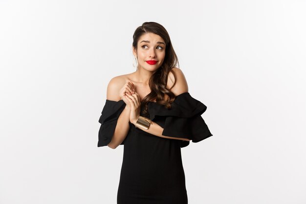 Fashion and beauty. Young silly woman in black dress pouting and looking timid, standing in black glamour dress over white background.