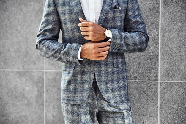 Модель бизнесмена моды одела в элегантном изменчивом костюме, позирующем около серой стены на фоне улицы. Метросексуал с роскошными часами на запястье