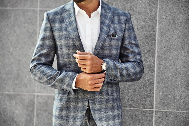 Fashion businessman model dressed in elegant checkered suit posing near gray wall on street background. Metrosexual with luxury watch on wrist