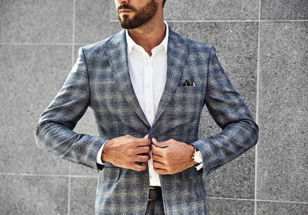 Fashion businessman model dressed in elegant checkered suit posing near gray wall on street background. Metrosexual with luxury watch on wrist