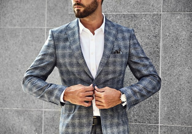 패션 사업가 모델 거리 배경에 회색 벽 근처 포즈 우아한 체크 무늬 양복을 입고. 손목에 고급 시계가있는 메트로 섹슈얼
