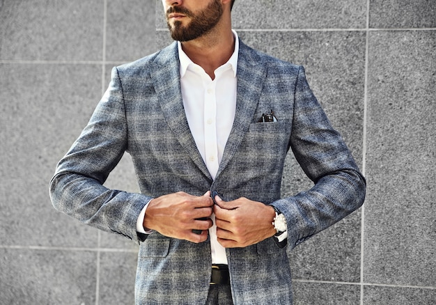 Fashion businessman model dressed in elegant checkered suit posing near gray wall on street background. Metrosexual with luxury watch on wrist