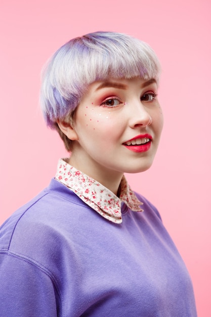 Мода портрет крупным планом улыбающейся красивой кукольной девушки с короткими светлыми фиолетовыми волосами в сиреневом свитере на розовой стене