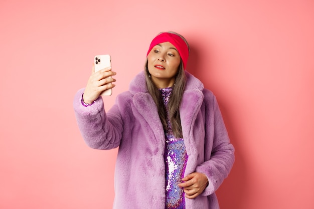 ファッションコンセプト。スタイリッシュなアジアのシニア女性がスマートフォンで自分撮りをし、紫色のフェイクファーのコートとパーティードレスでポーズをとって、ピンクの背景の上に立っています。