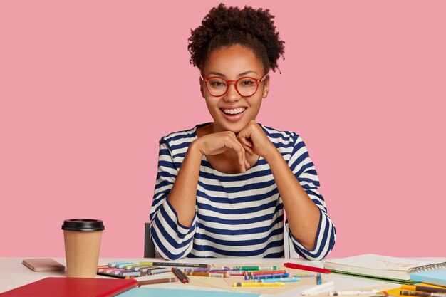 Fashion designer with toothy smile, feels sarisfied, keeps hands under chin, wears striped clothes, isolated over pink wall. Positive painter has inspiration for drawing. Creative work concept