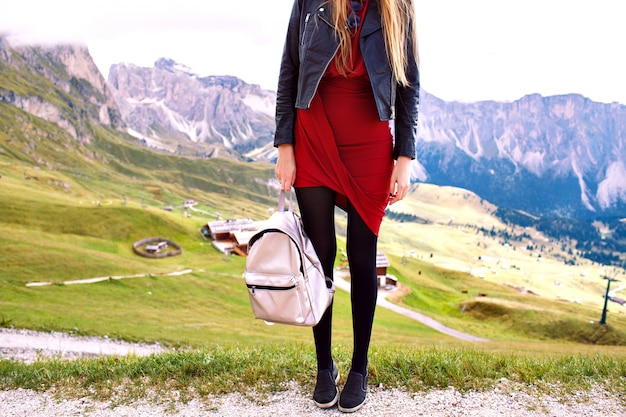 Fashion details of stylish tourist woman posing eating elegant dress leather jacket and trendy backpack, fashionable luxury vacation in Alp mountains.