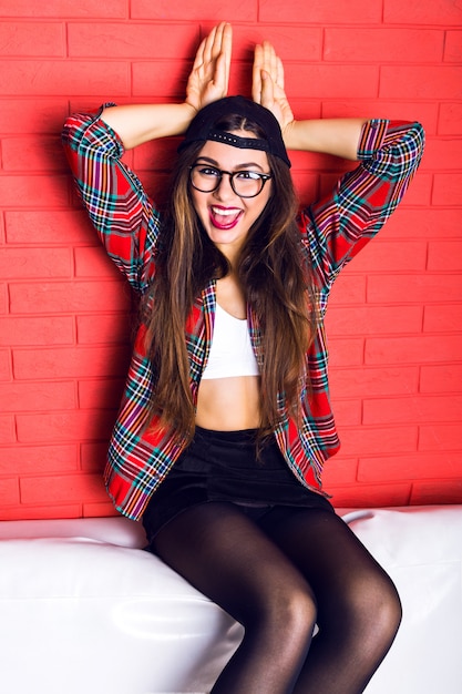 Fashion funny indoor portrait of young pretty hipster woman having fun alone at wed urban wall, have stunning long brunette hairs and bright make up, wearing glasses,swag hat, plaid shirt.