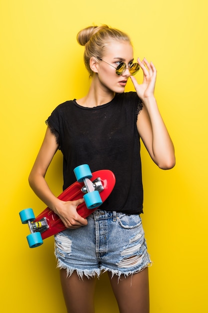 Moda ragazza cool hipster sorridente felice in occhiali da sole e vestiti colorati con skateboard divertendosi all'aperto sullo sfondo arancione
