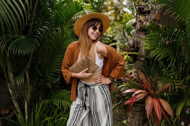 熱帯のヤシの葉でポーズ麦わら帽子のセクシーな優雅な女性のファッション画像