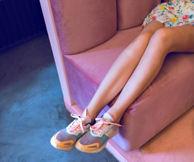 여자 긴 슬림 다리의 패션 이미지, 파스텔 스니커즈와 장미 소파에 앉아 미니 드레스, 네온 분위기, 부드러운 색상.