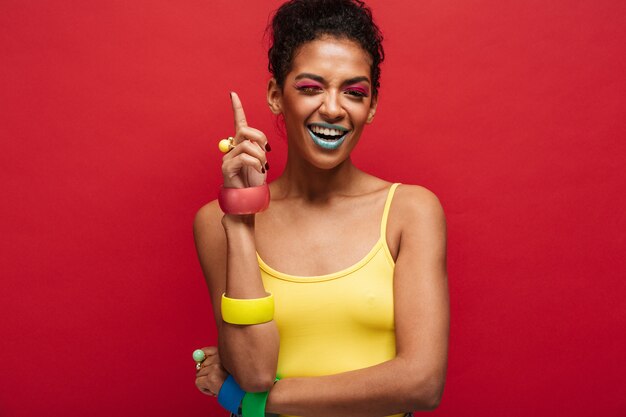 Fashion joyful african american female model in yellow shirt smiling and pointing finger upward, isolated over red wall