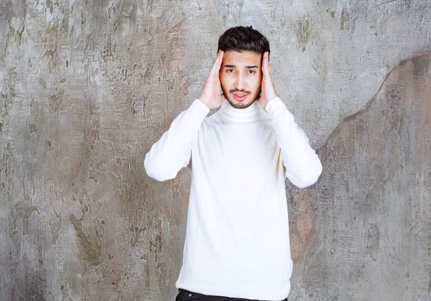 Fashion model in white sweater standing on concrete wall and looking confused or thoughtful.