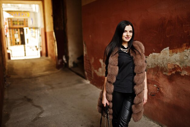 Fashion outdoor photo of gorgeous sensual woman with dark hair in elegant clothes and luxurious fur coat at old street with grunge walls