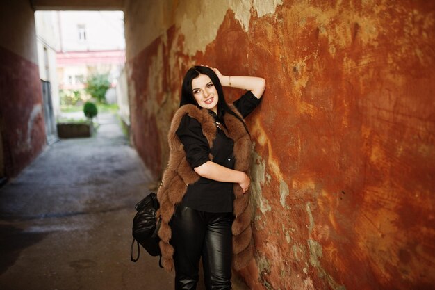 Fashion outdoor photo of gorgeous sensual woman with dark hair in elegant clothes and luxurious fur coat at old street with grunge walls