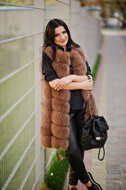 Fashion outdoor photo of gorgeous sensual woman with dark hair in elegant clothes and luxurious fur coat and with backpack against iron fence