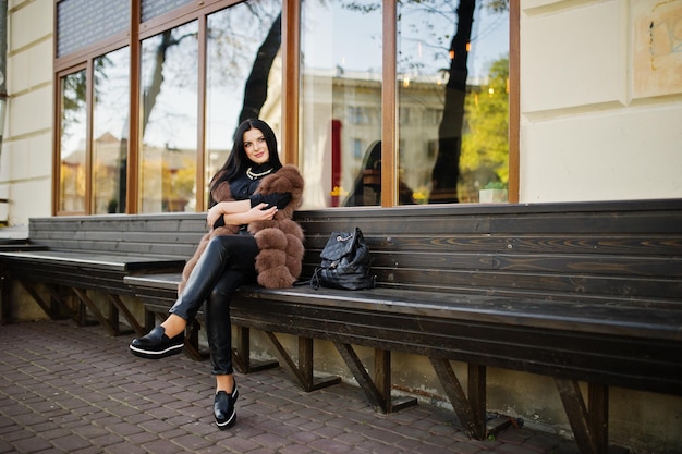 Fashion outdoor photo of gorgeous sensual woman with dark hair in elegant clothes and luxurious sleeveless fur coat and with backpack sitting on bench at autumn city
