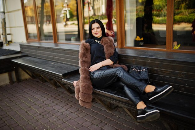Fashion outdoor photo of gorgeous sensual woman with dark hair in elegant clothes and luxurious sleeveless fur coat and with backpack sitting on bench at autumn city