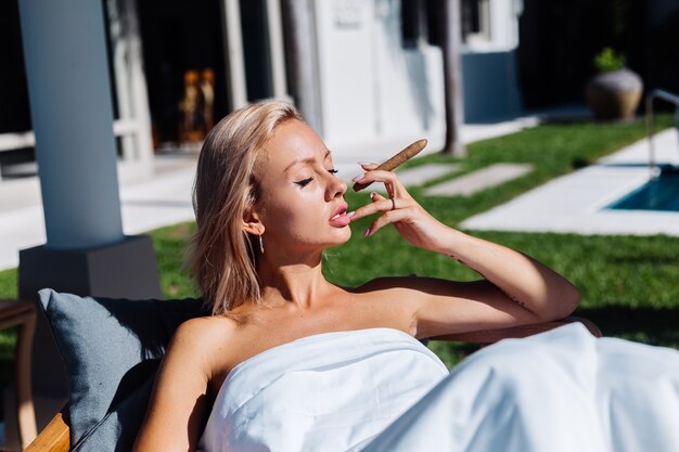 Модный открытый портрет обнаженной женщины сидит на диване, накрываясь одеялом, держа сигару