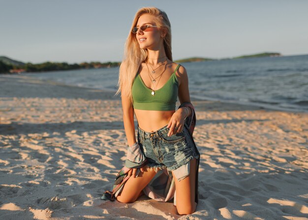 Fashion photo of sexy  blond woman in green crop top and jeans posing on tropical beach.