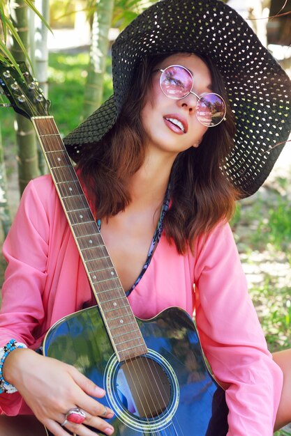 自然なメイクアップとギターで庭でポーズをとってふわふわのブルネットの毛の美しい少女のファッションの肖像画。帽子と丸いトレンディなピンクのサングラス。