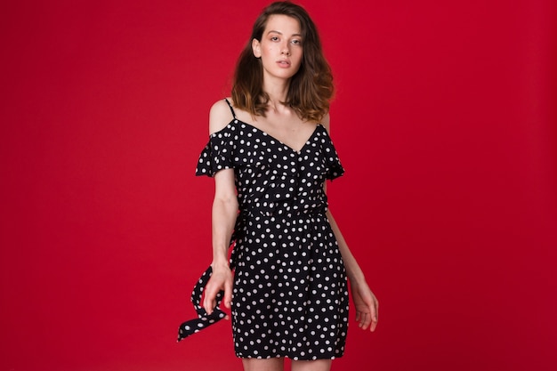 Fashion portrait of beautiful young woman in black dotted dress on red studio