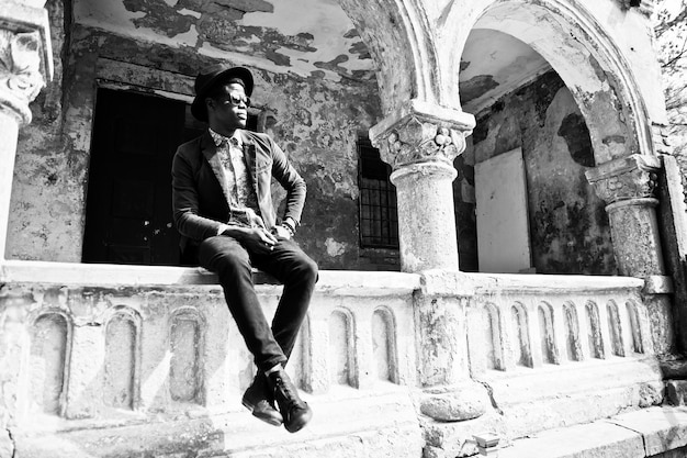 Fashion portrait of black african american man on green velvet jacket and black hat sitting on the railing background old vintage house with arch BW photo