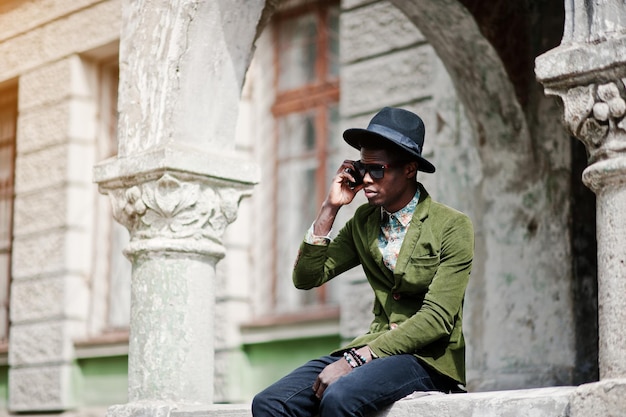 Fashion portrait of black african american man on green velvet jacket and black hat sitting on the railing and talking on his cell phone background old vintage house with arch