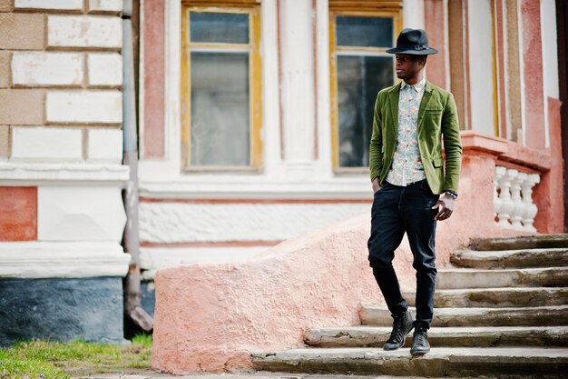 Fashion portrait of black african american man on green velvet jacket and black hat stay on stairs background old mansion