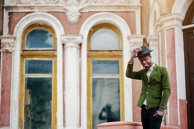 Fashion portrait of black african american man on green velvet jacket stay on stairs and wear his black hat background old mansion