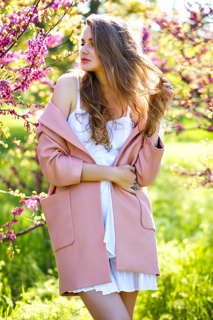 Fashion portrait of blissful stunning elegant woman posing at park with blooming Sakura trees at spring time