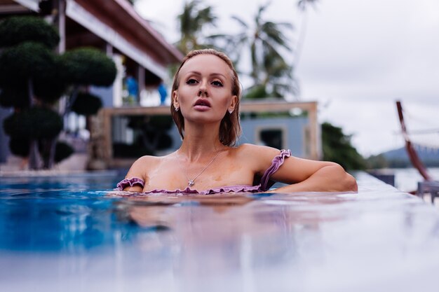 Fashion portrait of caucasian woman in bikini in blue swimming pool on vacation at coudy day natural light