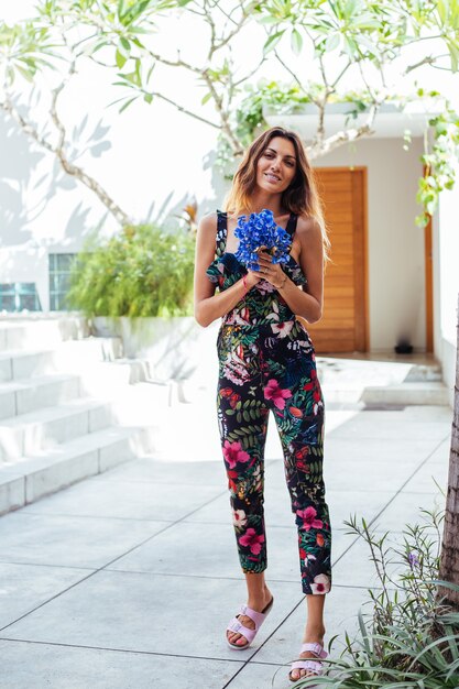 Fashion portrait of caucasian woman in stylish summer jumpsuit with flowers outside villa