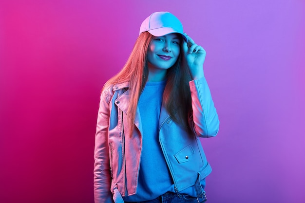 Fashion portrait of confident woman standing against pink neon wall and touching visor of her cap, wearing leather jacket, looking at camera