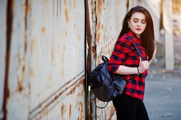 Fashion portrait girl with red lips wearing a red checkered shirt and backpack background rusty fence