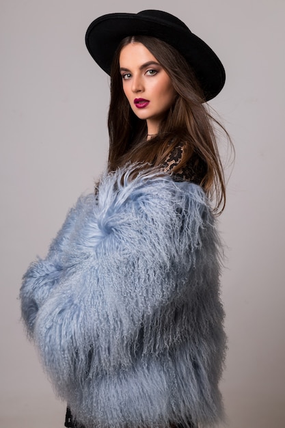 fashion portrait of gorgeous  woman in stylish winter fluffy  blue coat and black hat posing on bright  grey wall.