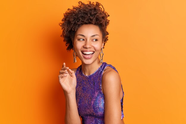 Fashion portrait of happy stylish woman in bright clothes earrings smiles broadly looks aside positively isolated over orange wall