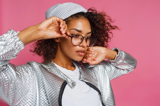 Fashion portrait of  mix race woman with brown skin and curly African hairstyle on vivid pink background. Wearing silver winter  jacket and grey hat.