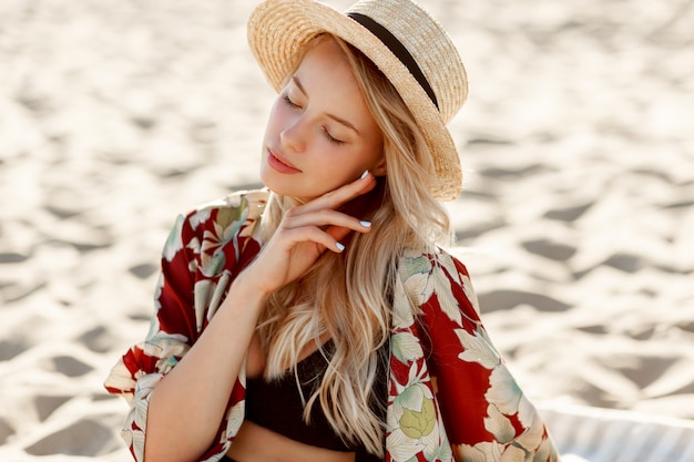 太陽が降り注ぐビーチで休んでいる自然なメイクでゴージャスな金髪の女性のファッションの肖像画。麦わら帽子をかぶっています。休日と休暇の気分。